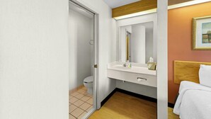 A hotel bathroom with a toilet, sink, mirror, and a bed with a wooden headboard.