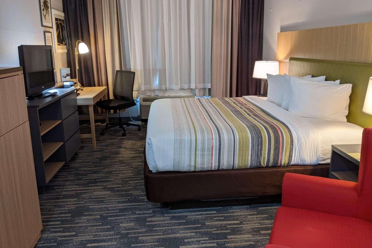A hotel room with a bed, a desk with a chair, a television, and a red armchair.