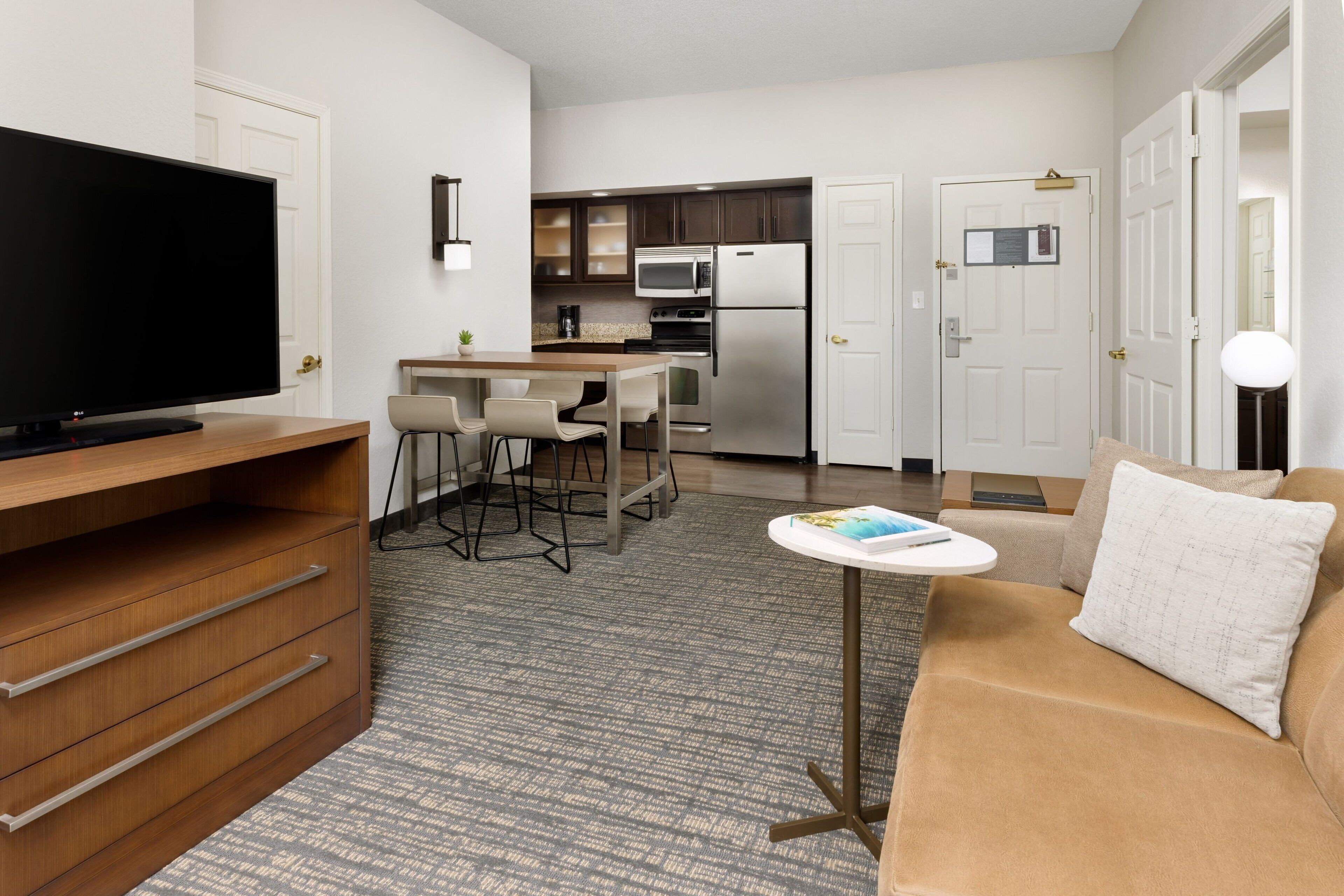 A modern hotel room with a flat-screen TV, a sofa, a small table, a kitchen area with a microwave and stove, and a carpeted floor.