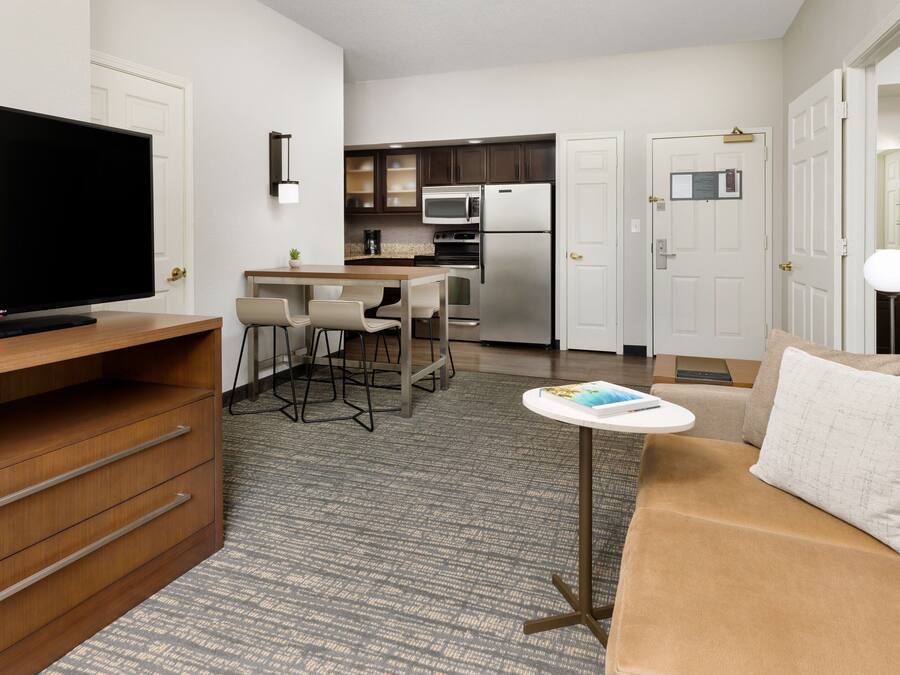 A modern hotel room with a flat-screen TV, a sofa, a small table, a kitchen area with a microwave and stove, and a carpeted floor.