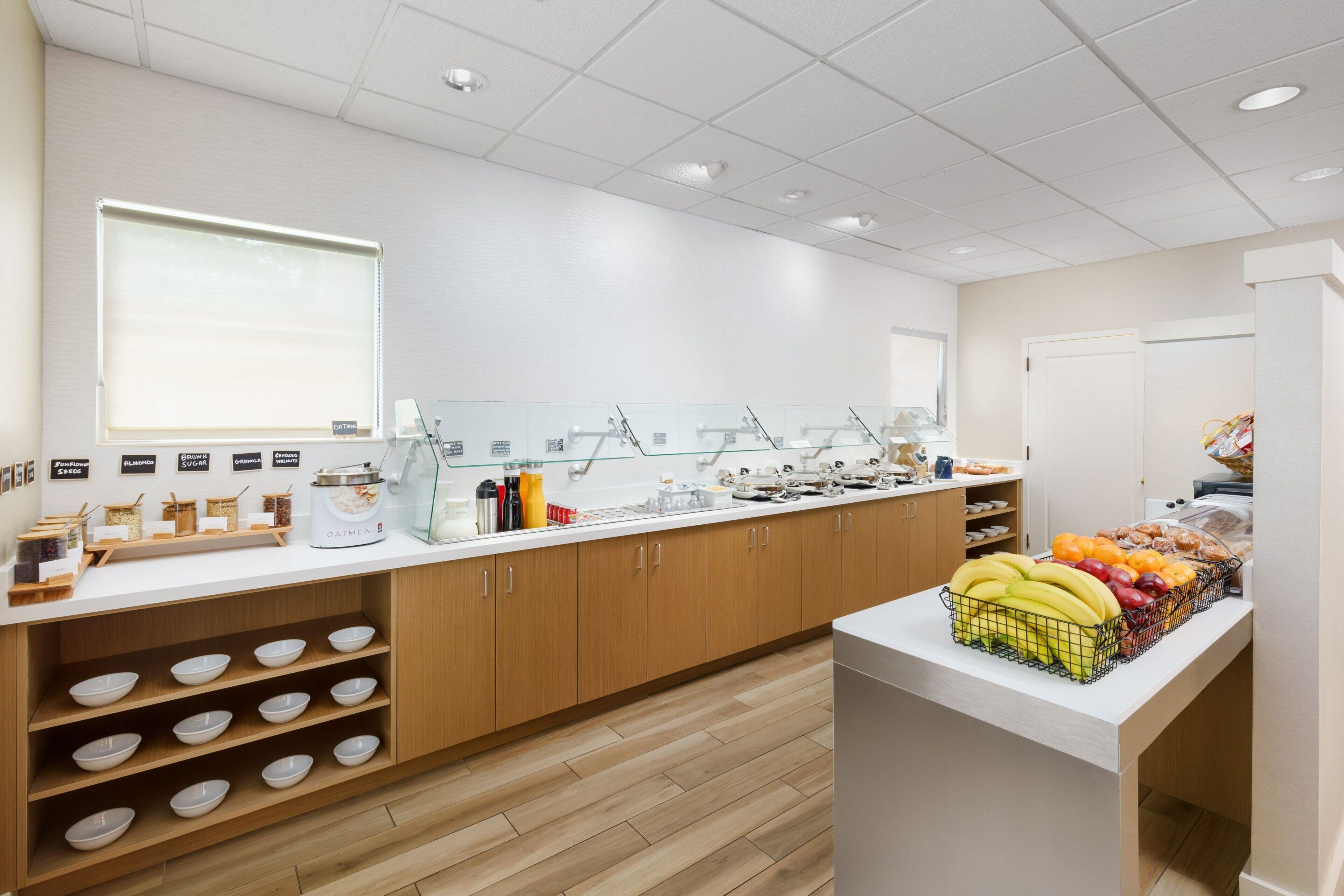 A clean, modern breakfast area with a white countertop, wooden cabinets, and a variety of food items including fruits and cereals.