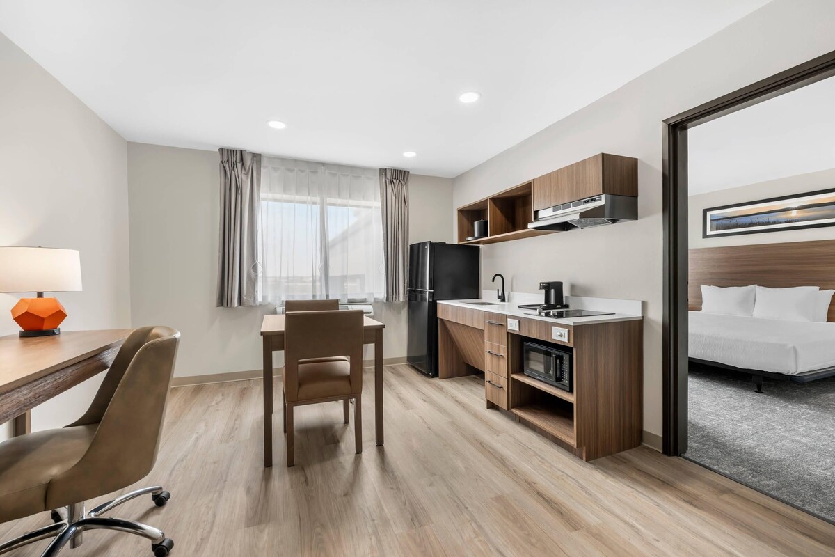 A hotel room with a bed, a desk with a chair, a kitchenette, and a window with curtains.