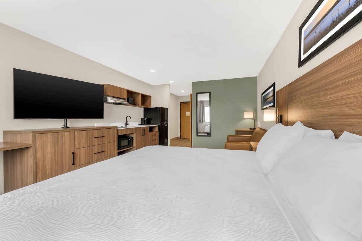 Une chambre d’hôtel moderne dotée d’un grand lit, d’un téléviseur à écran plat, d’armoires en bois, d’une kitchenette et donnant sur le couloir.