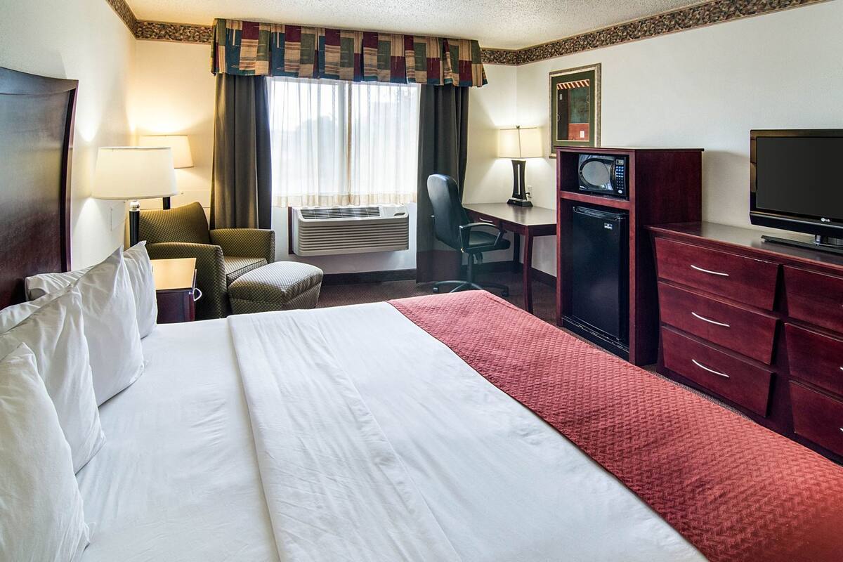 A hotel room with a bed, a desk with a computer, a TV, an air conditioning unit, and a chair.