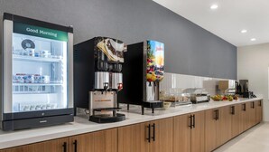 A modern kitchen area with a refrigerator, coffee machine, juice dispenser, and a counter with various food items.