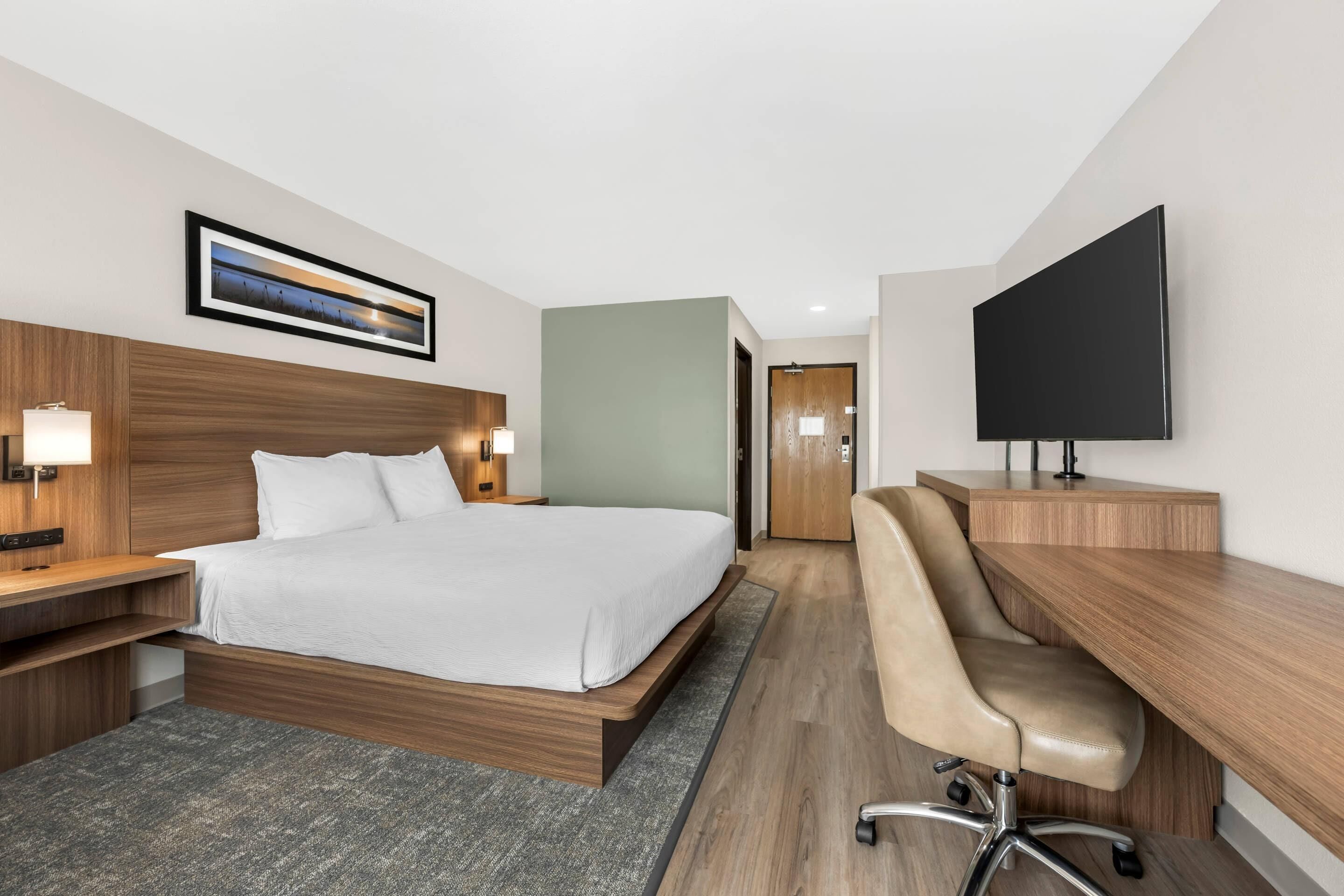 A hotel room with a bed, desk, chair, TV, and artwork.