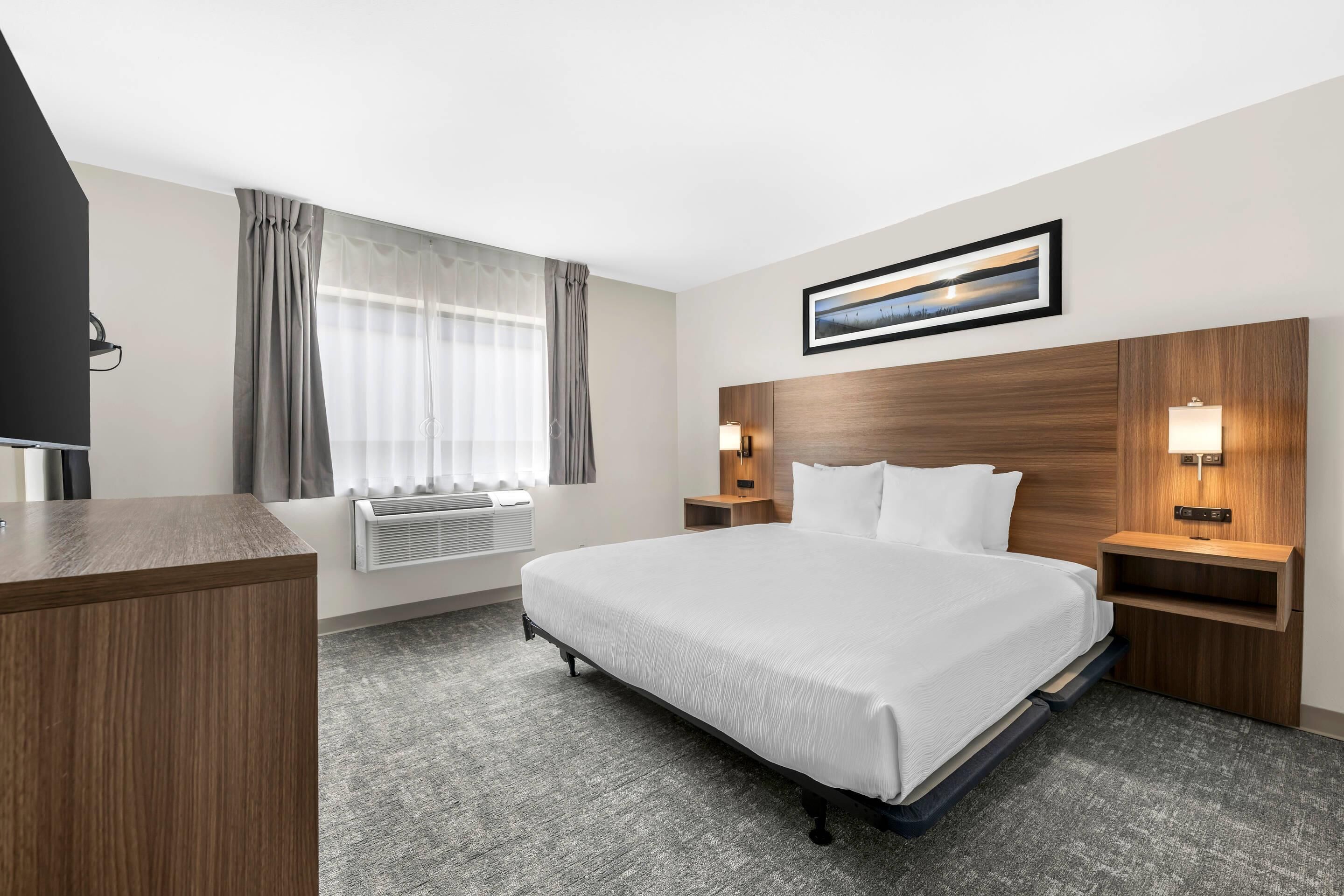 A hotel room with a large bed, a desk, a TV, a window with curtains, a framed picture on the wall, and two bedside lamps.