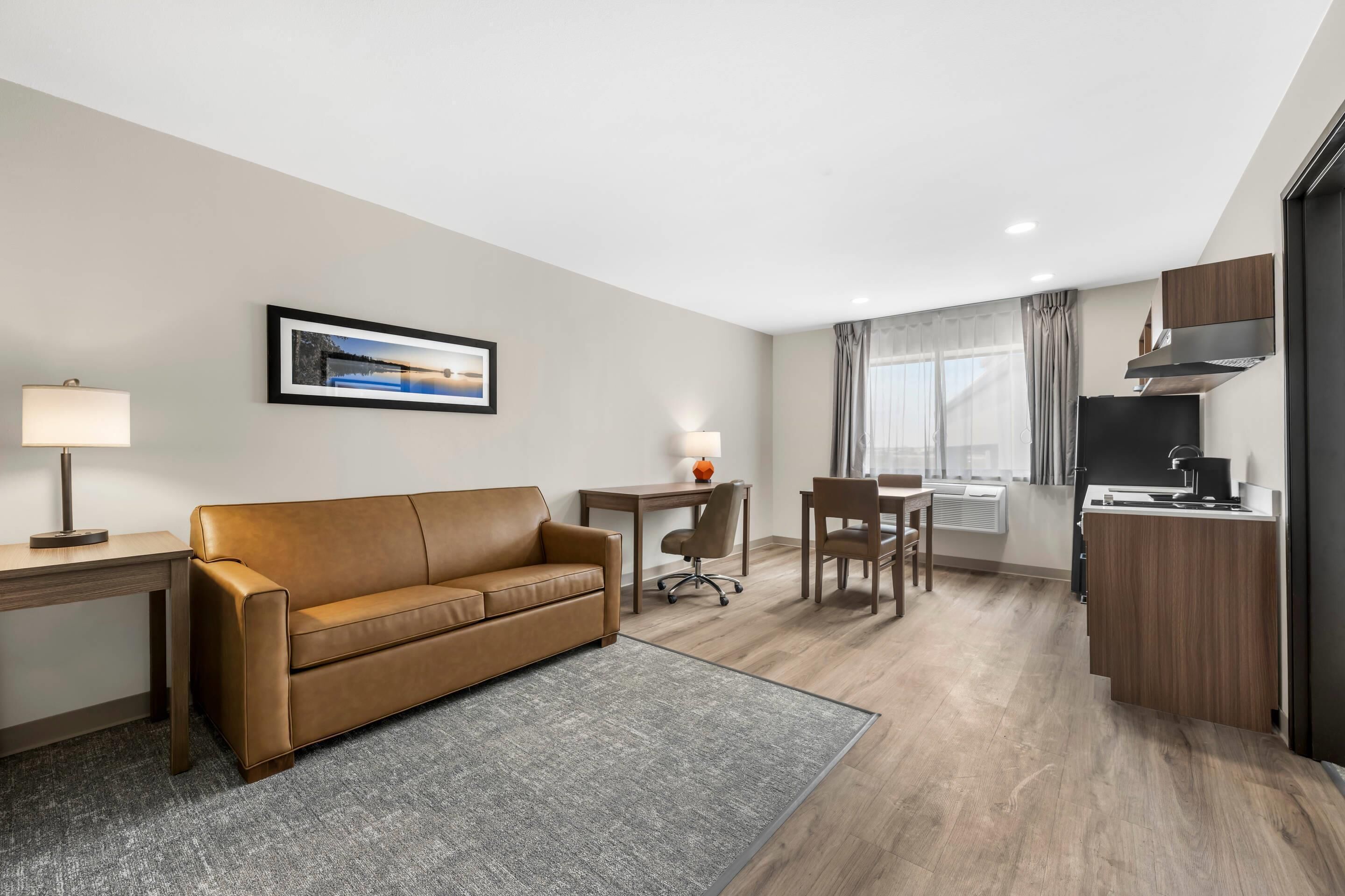 A modern hotel room with a leather sofa, a dining table, a kitchen area, and a framed picture on the wall.