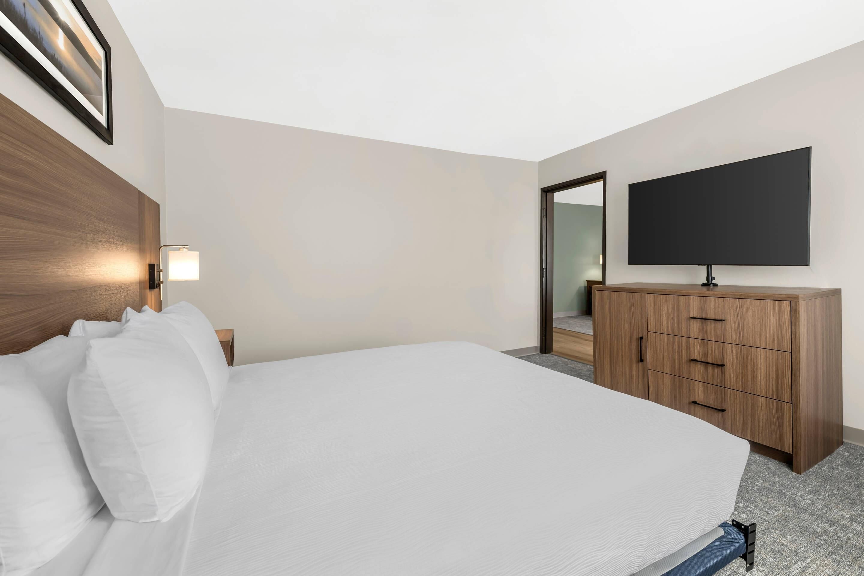 Une chambre d’hôtel dotée d’un grand lit, d’une tête de lit en bois, d’un téléviseur à écran plat fixé au mur et d’une commode en bois avec des tiroirs.