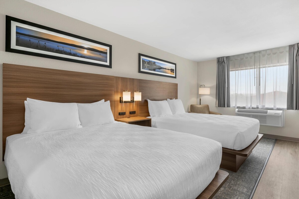Une chambre d’hôtel avec deux lits, une tête de lit en bois, des lampes fixées au mur, une fenêtre avec des rideaux et des œuvres d’art encadrées accrochées au mur.
