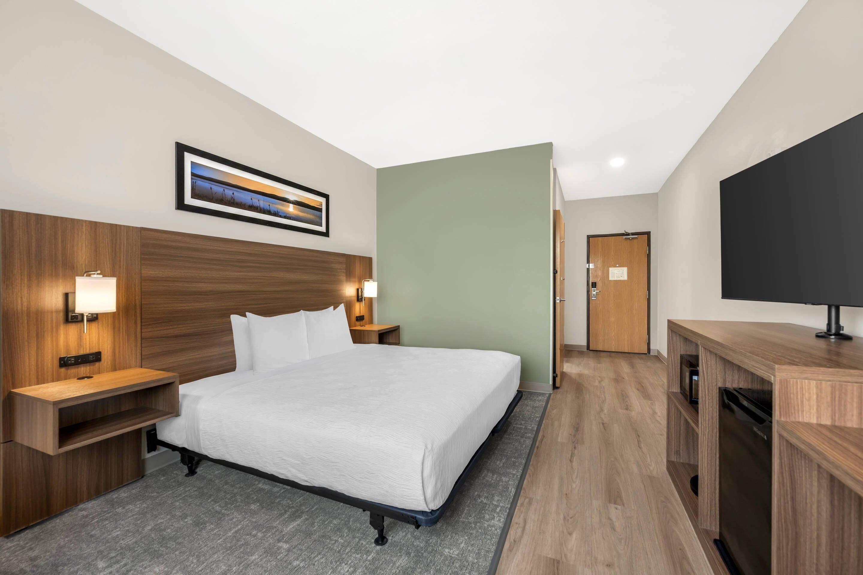 Habitación de hotel con cama, lámparas de noche, televisor de pantalla plana, cabecero de madera, un cuadro enmarco y una puerta.