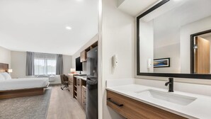 Une chambre d’hôtel moderne équipée d’un lit, d’un bureau, d’un réfrigérateur, d’un grand miroir et d’une salle de bain avec un lavabo et un miroir.