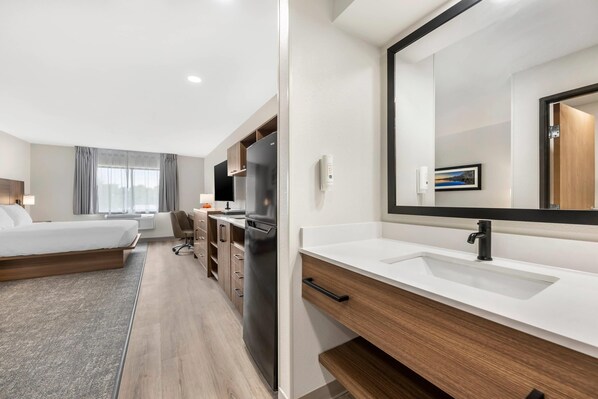 Una habitación de hotel moderna con cama, escritorio, refrigerador, un espejo grande y un baño con lavamanos y espejo.
