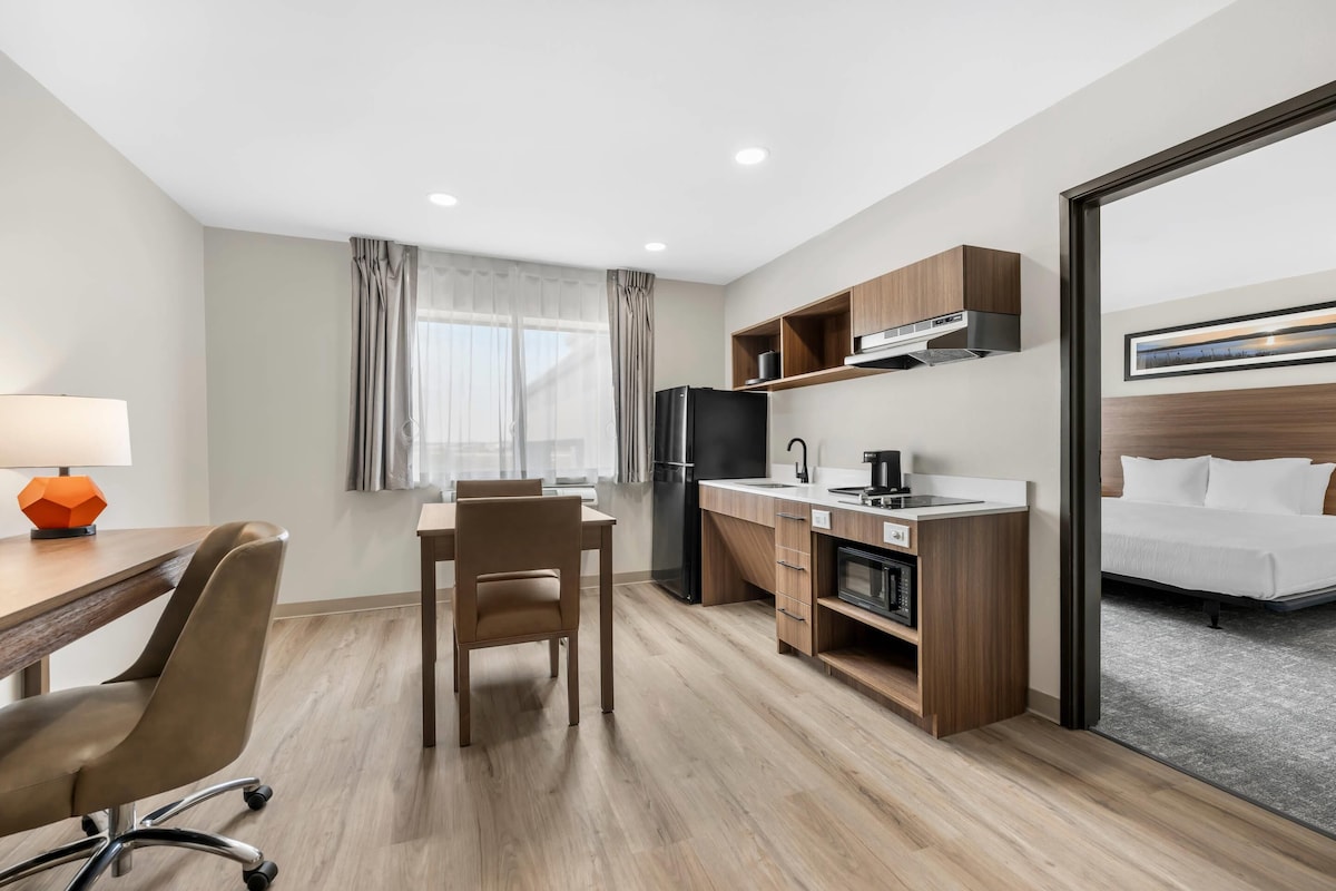 A hotel room with a bed, a desk with a chair, a kitchenette, and a window with curtains.