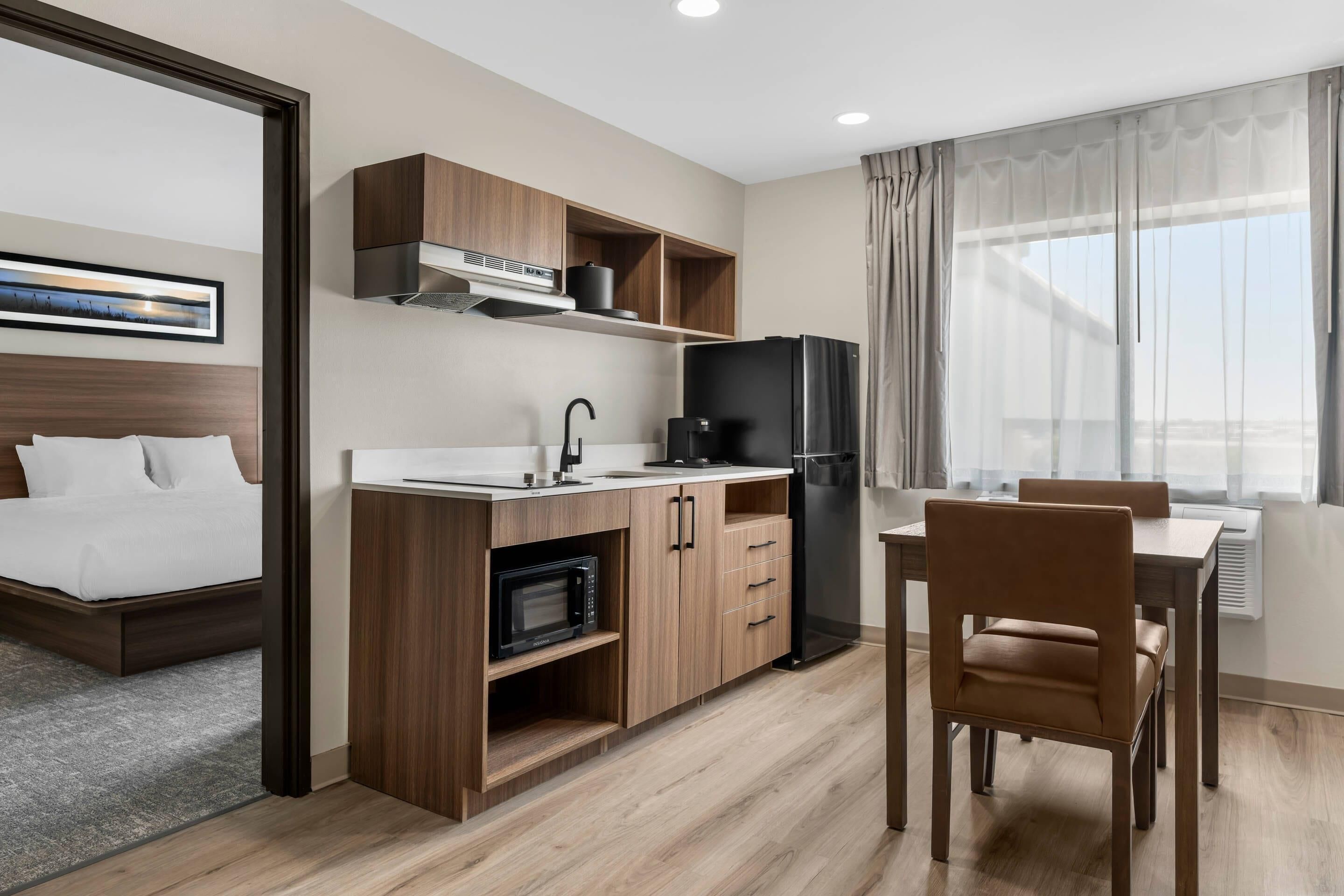 Habitación de hotel compacta con kitchenette, cama, comedor y un ventanal grande con cortinas.