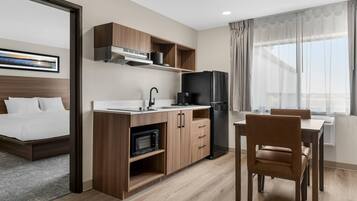 Habitación de hotel compacta con kitchenette, cama, comedor y un ventanal grande con cortinas.