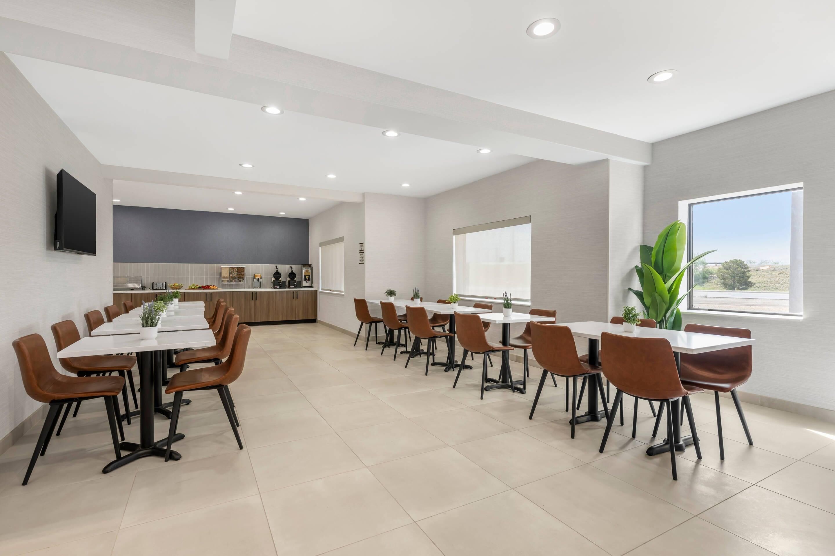 Un comedor moderno con varias mesas y sillas, una cocina al fondo y un amplio ventanal que aporta luz natural.