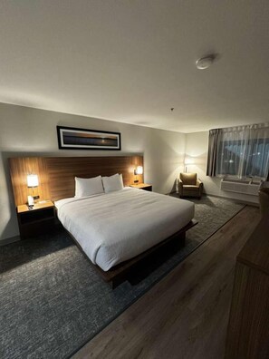Quarto de hotel com uma cama grande, duas mesas de cabeceira com abajures, uma cadeira, uma janela com cortinas e um quadro na parede.