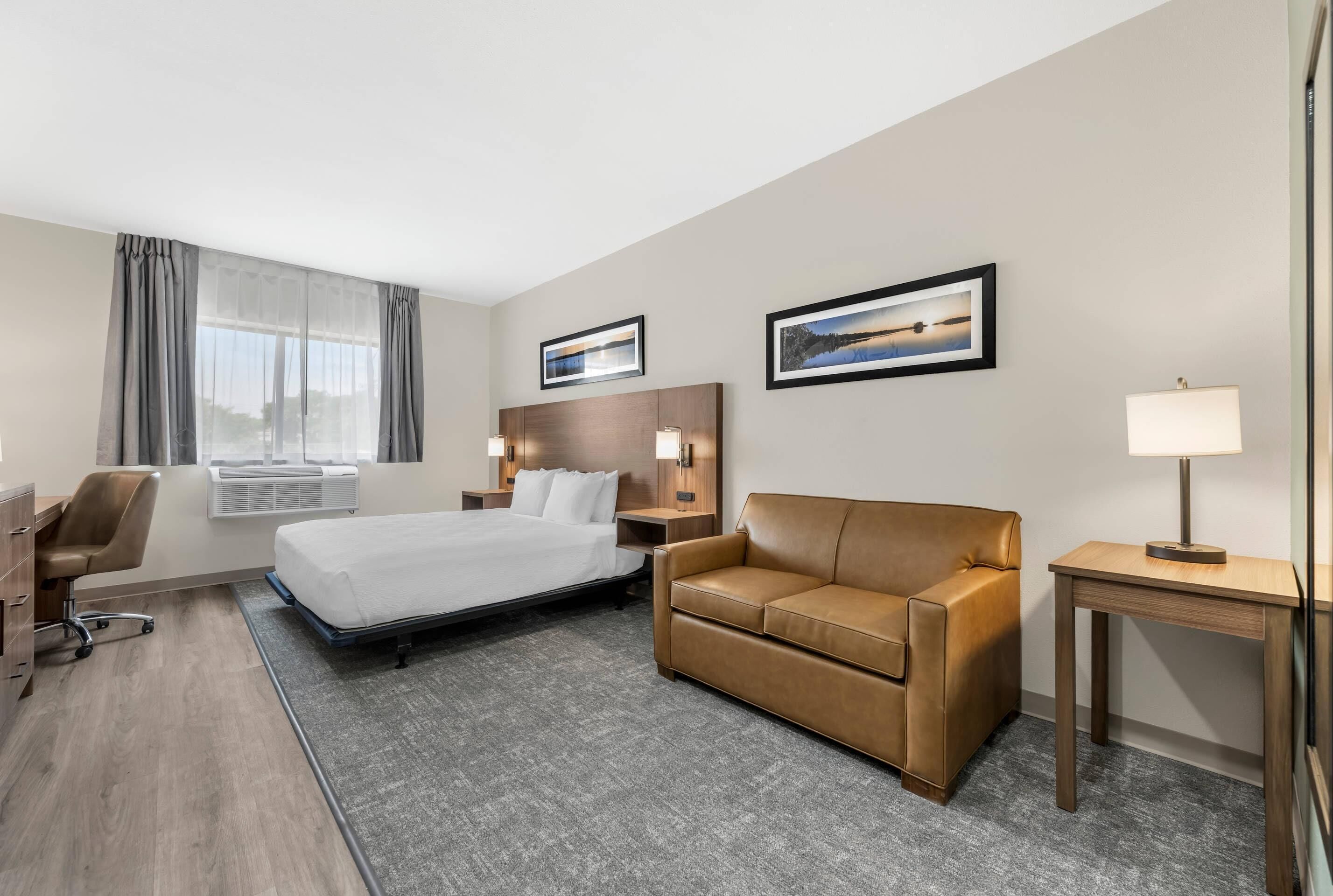 Une chambre d’hôtel avec un lit, un canapé en cuir marron, un bureau avec une chaise, une table de chevet avec une lampe, et deux tableaux encadrés au mur.