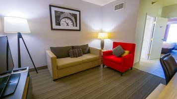 Una habitación de hotel con un sofá, un sillón rojo, una lámpara, un reloj en un marco y una cama al fondo.