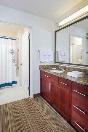 A hotel bathroom with a double sink, a large mirror, a shower with a blue and white curtain, and a wooden floor. - Residence Inn Waco (Waco)