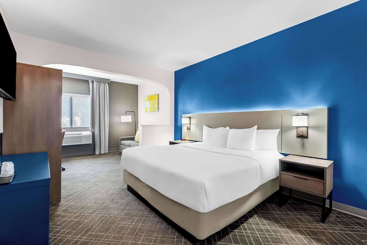 A hotel room with a large bed, a nightstand with a lamp, a desk with a chair, and a window with curtains.