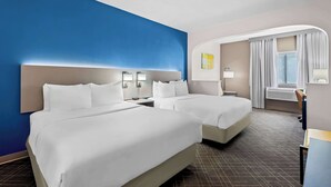 A hotel room with two beds, a desk, a chair, a window with curtains, and a wall with a blue accent.