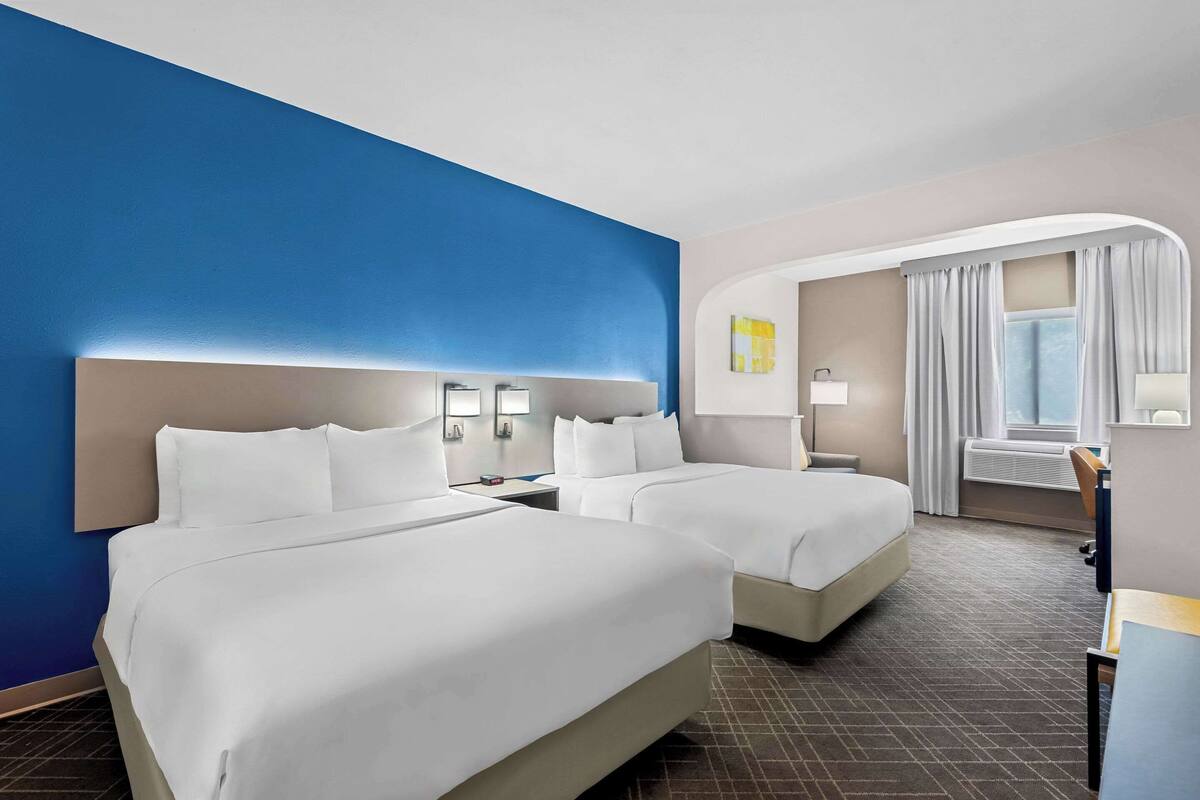 A hotel room with two beds, a desk, a chair, a window with curtains, and a wall with a blue accent.
