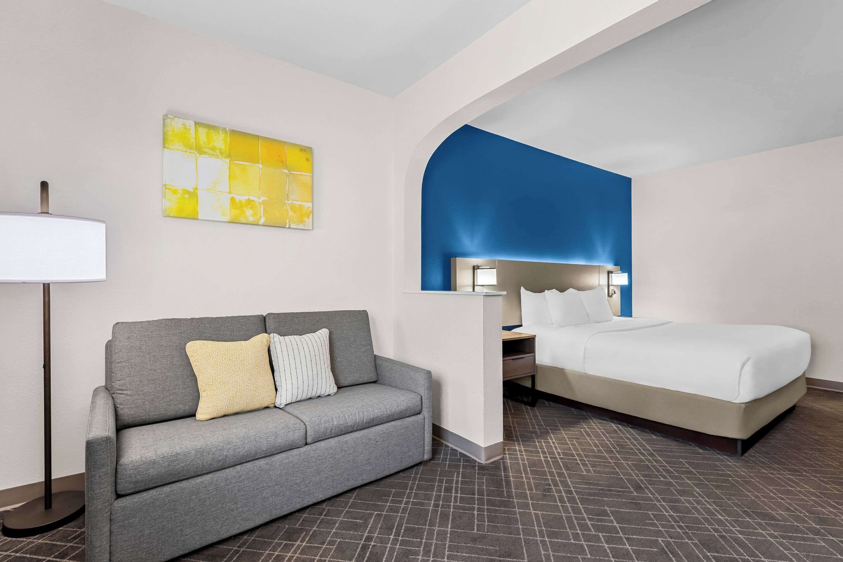 Een hotelkamer met een grijze bank, een wit bed, een bureau met een lamp en een blauwe accentmuur.