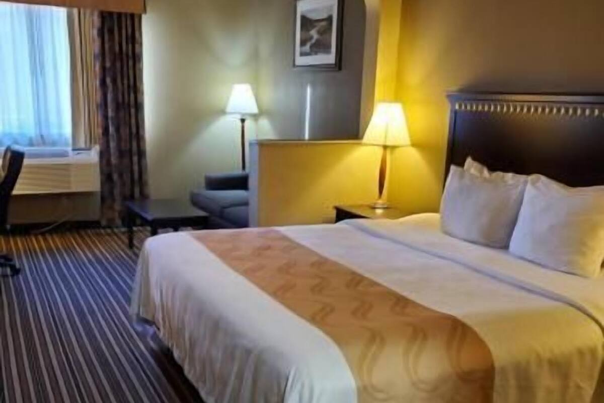 A hotel room with a large bed, a desk, a chair, a lamp, and a window with curtains.