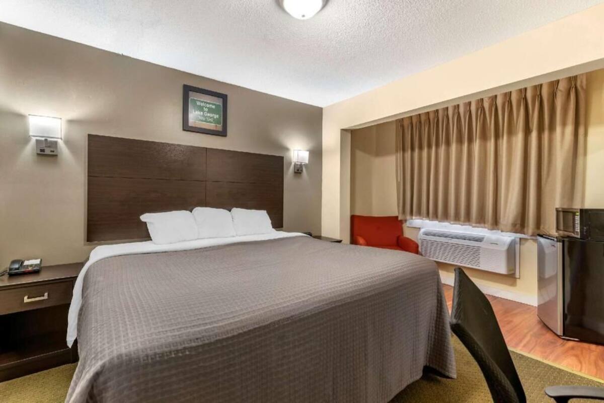A hotel room with a bed, bedside table, lamp, air conditioning unit, and a red chair.