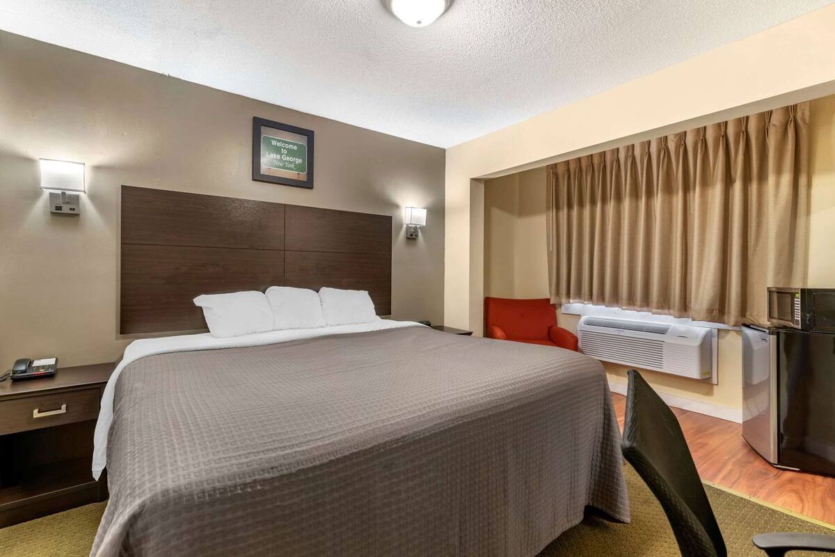 A hotel room with a bed, bedside table, lamp, air conditioning unit, and a red chair.
