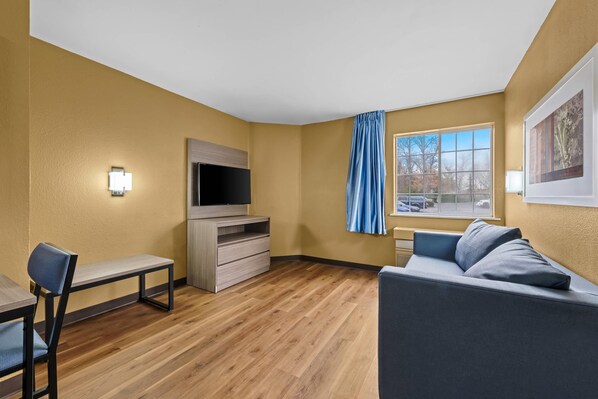 A modern hotel room with a wooden floor, a gray sofa, a small table, a TV, and a window with blue curtains. - Suburban Studios Charlotte – Executive Park (Charlotte)