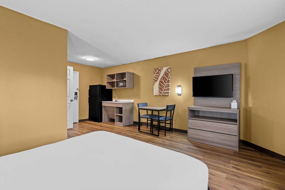 Habitación de hotel con una cama, un escritorio con silla, televisor, armario y estante en la pared.