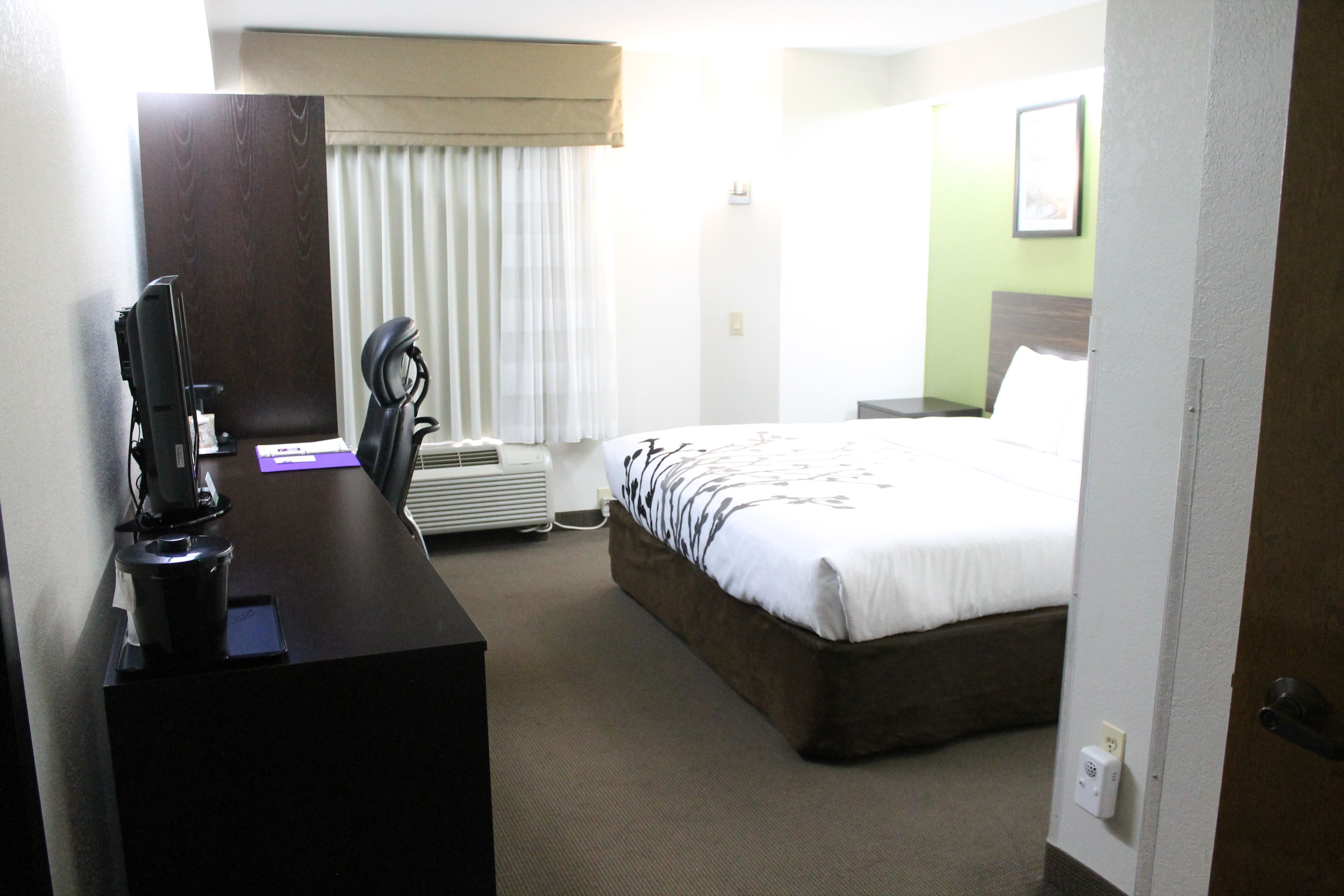 A hotel room with a bed, a desk with a computer, a chair, a window with curtains, and a wall-mounted thermostat.