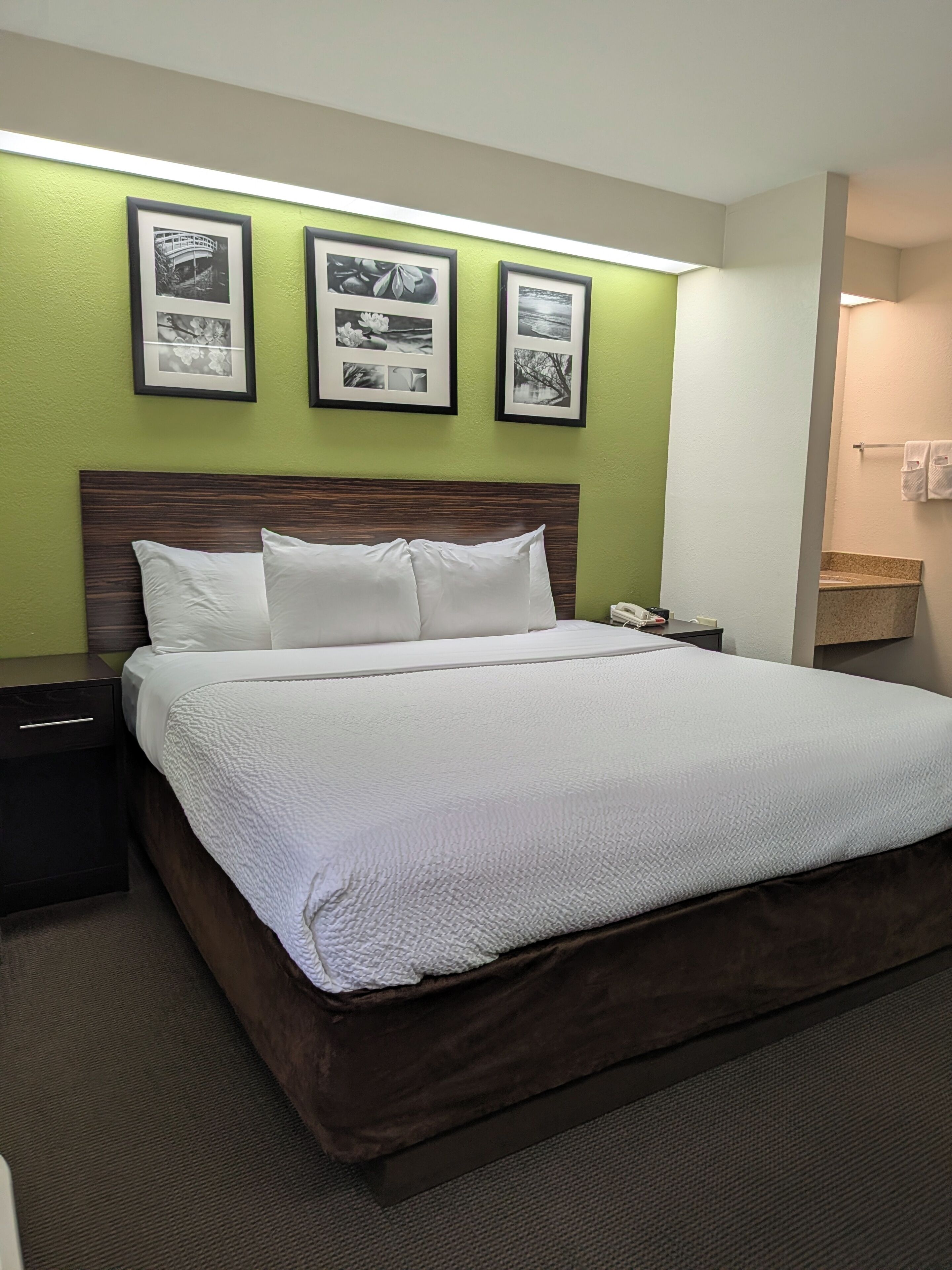 A hotel room with a large bed, a nightstand, a wall-mounted phone, and framed pictures above the bed.
