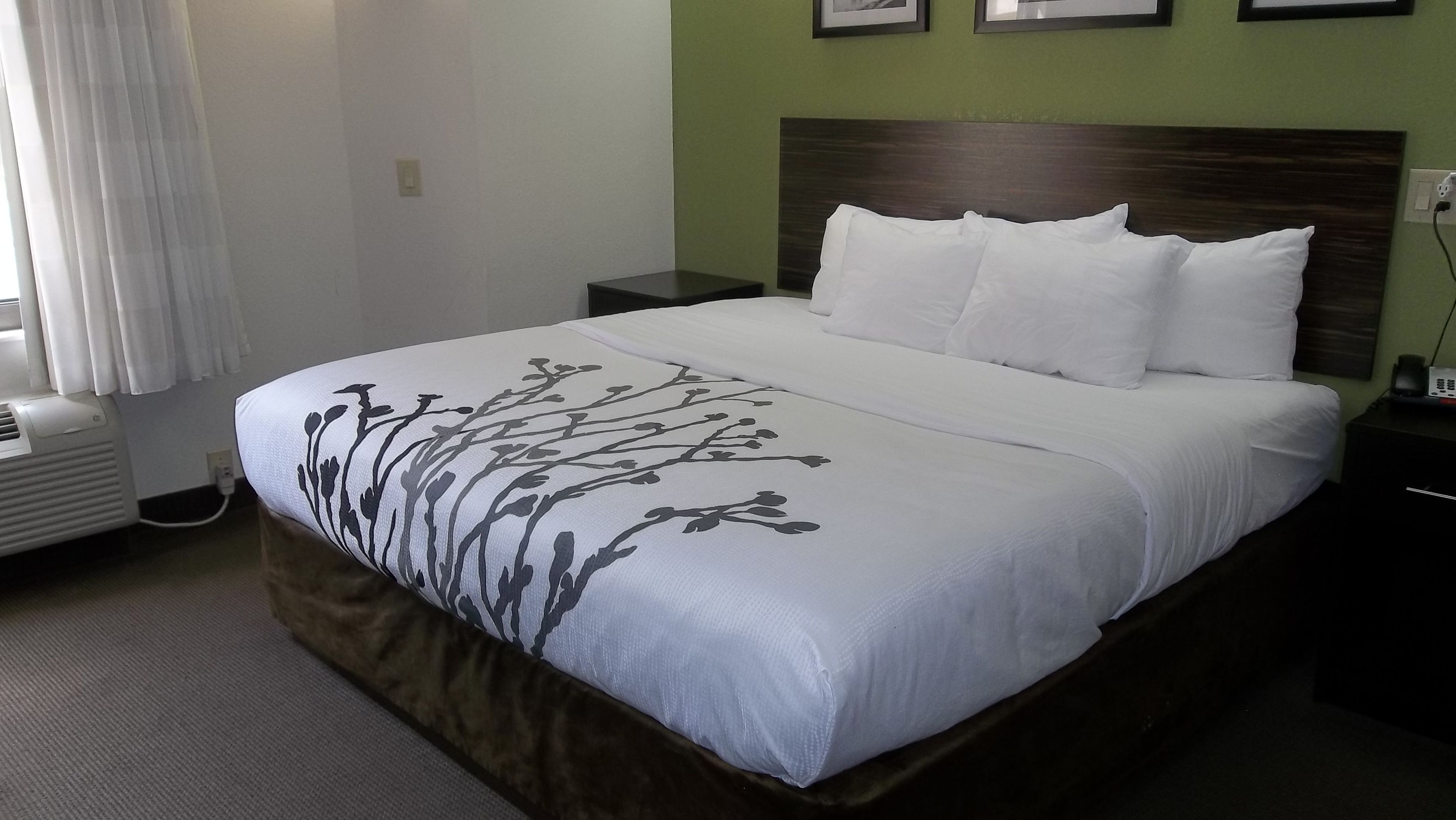 Un lit bien fait, recouvert d’une couvre-lit à motifs floraux noirs et blancs, avec une tête de lit sombre et une petite table de chevet sur laquelle se trouve un téléphone.