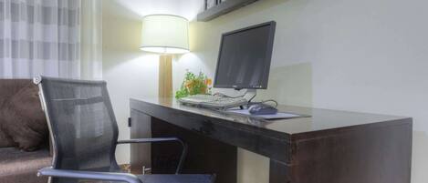 A hotel room with a desk, computer, chair, lamp, and a couch.