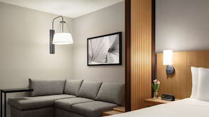 Ein Hotelzimmer mit Ecksofa, einem Bett, einem Nachttisch mit Lampe, einem gerahmten abstrakten Kunstwerk und einer Wandpaneelenverkleidung aus Holz.
