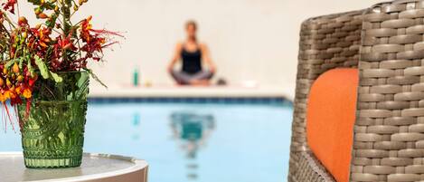 A wicker chair with an orange cushion, a green glass vase with flowers, and a person meditating by a pool.