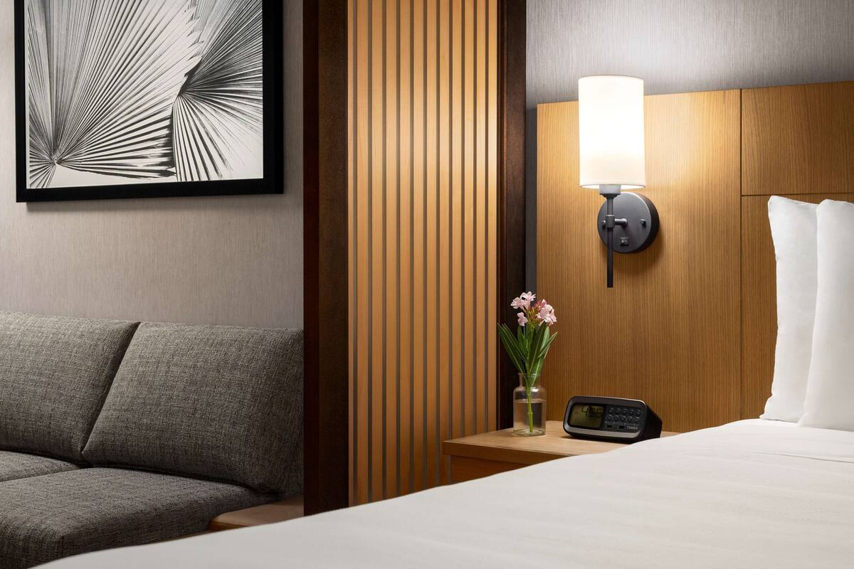 A hotel room with a bed, a nightstand with a lamp, a vase with flowers, and a framed picture on the wall.