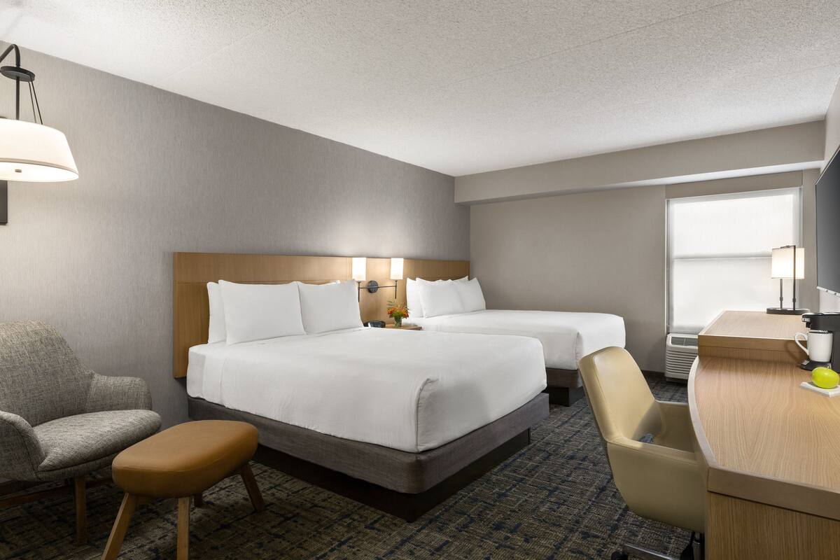 A hotel room with two beds, a desk, a chair, and a small stool.