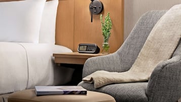 A hotel room with a bed, a gray armchair, a bedside table with a lamp, a small plant, and a book.