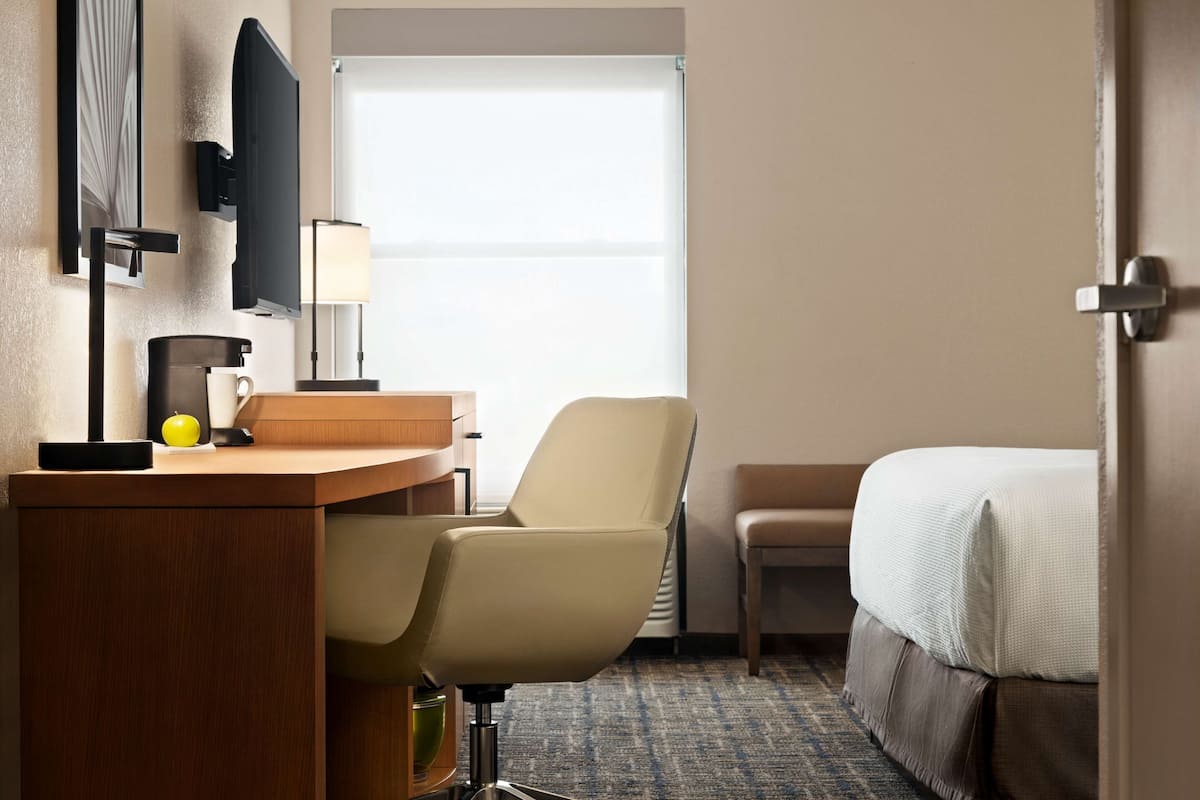 A hotel room with a bed, a desk with a chair, a TV mounted on the wall, and a window with blinds.
