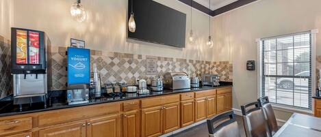 A modern kitchen with wooden cabinets, a black countertop, and a breakfast bar. There are coffee machines, a toaster, and a sign indicating breakfast is available for guests.
