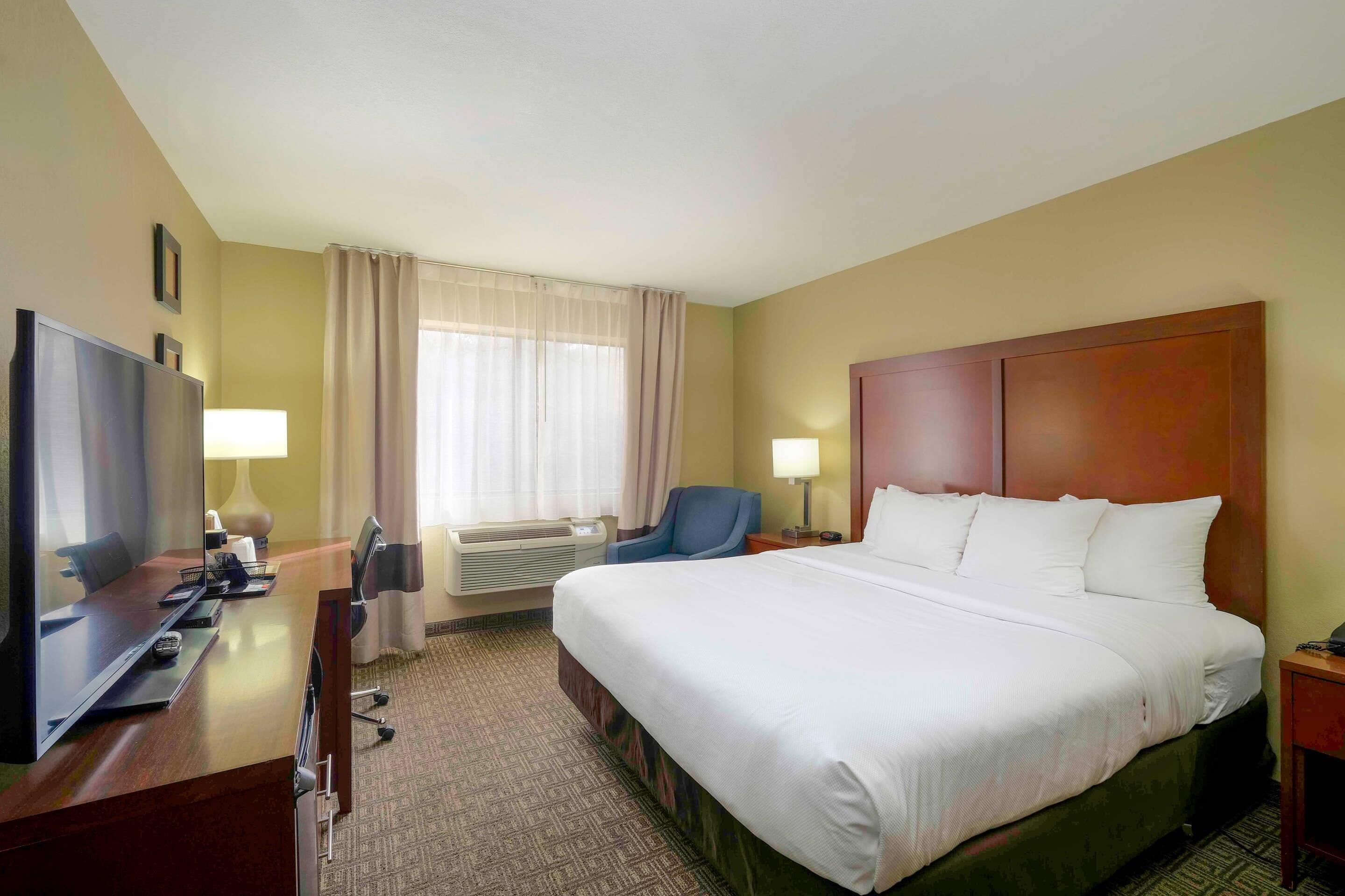 A hotel room with a large bed, a desk with a television, a chair, and a window with curtains.