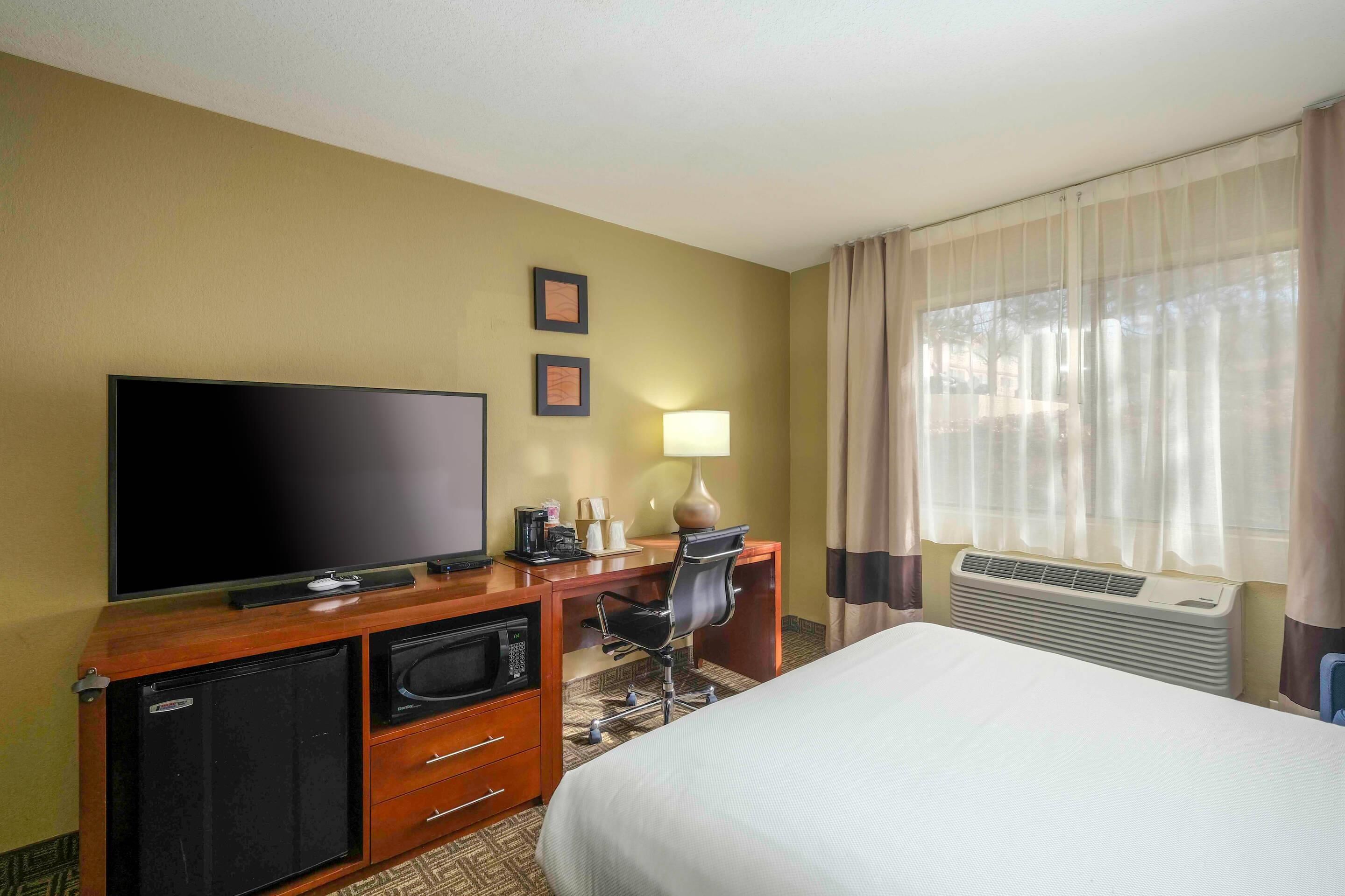 A hotel room with a bed, a desk with a flat-screen TV, a coffee maker, a lamp, and an air conditioner.