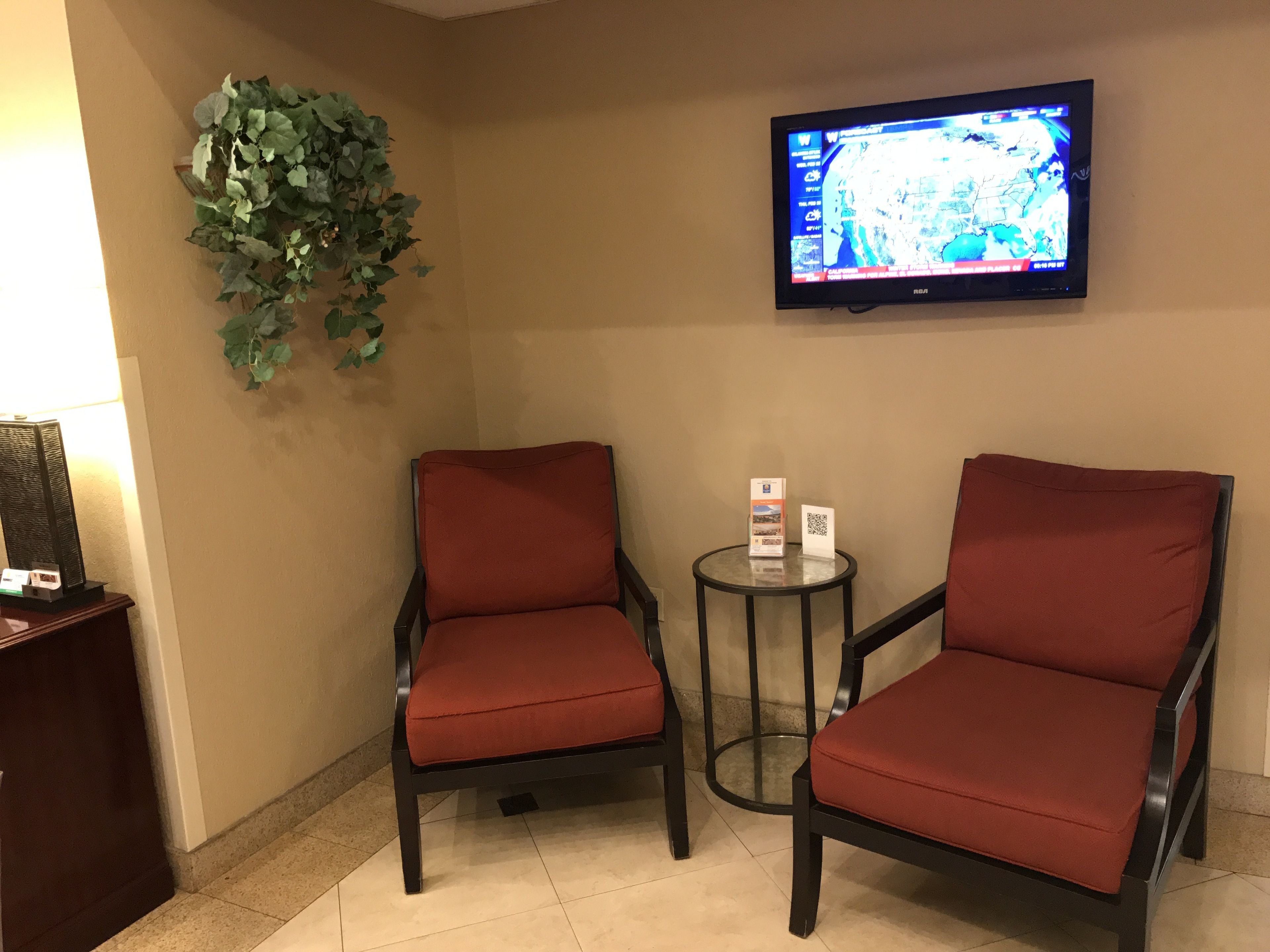 Two red chairs with black frames, a small round table with brochures, a television mounted on the wall displaying a map, and a potted plant on the left.