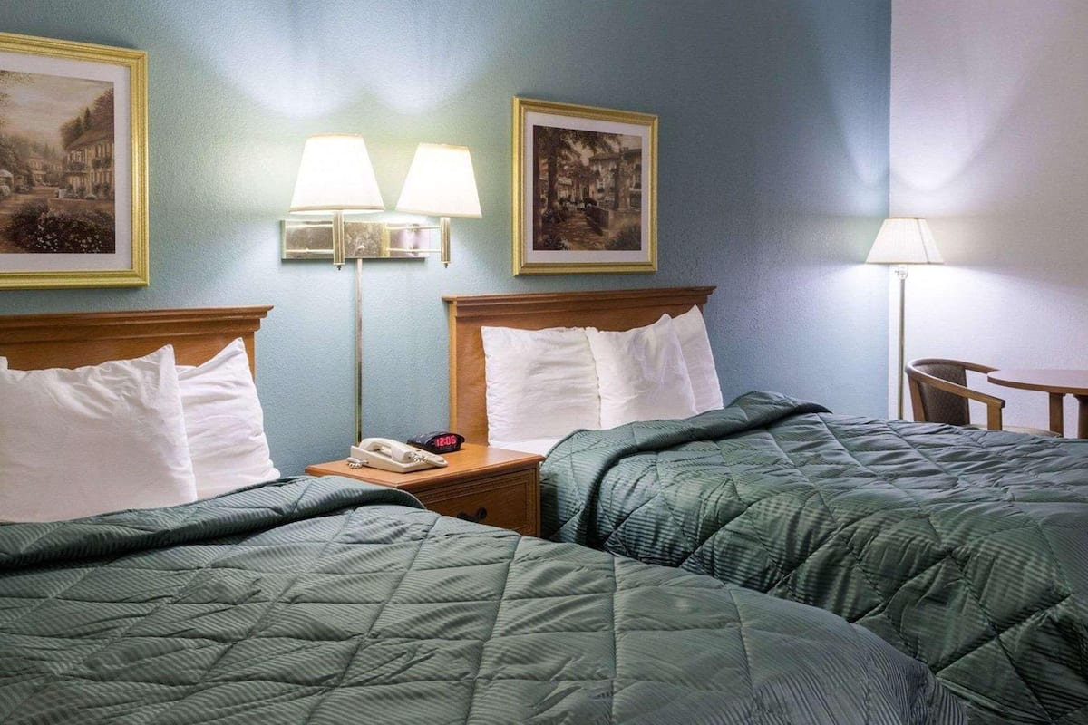 A hotel room with two beds, a nightstand with a phone and clock, and two wall-mounted lamps.