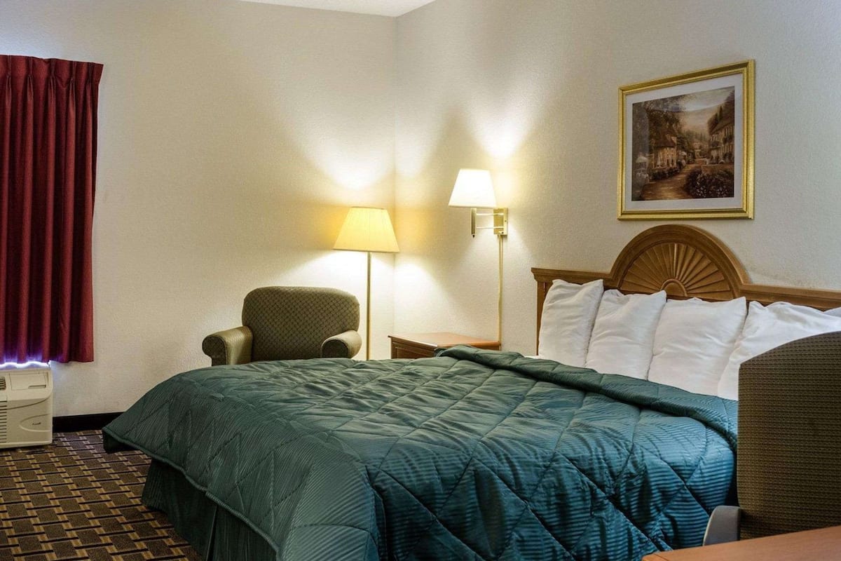 A hotel room with a bed, a chair, a desk, a lamp, and a painting on the wall.